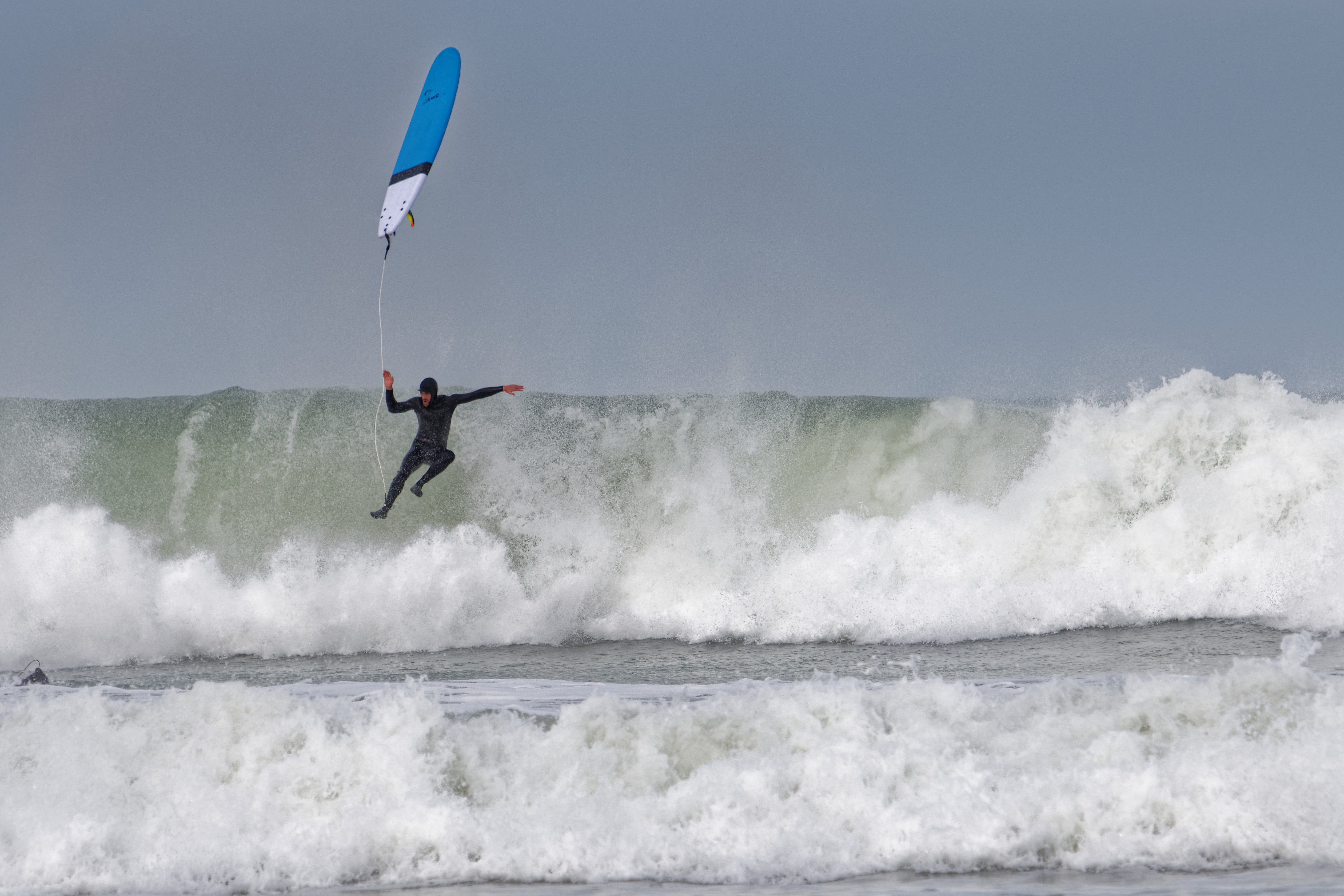 Alt text: "Un surfeur en combinaison noire fait un wipeout spectaculaire sur une vague massive. Sa planche bleue et blanche est projetée en l'air, reliée à sa cheville par le leash, tandis qu'il est en suspension dans les airs avec les bras écartés. La vague en arrière-plan est imposante avec de l'écume blanche, et l'eau présente une teinte vert-gris caractéristique des conditions de surf puissantes