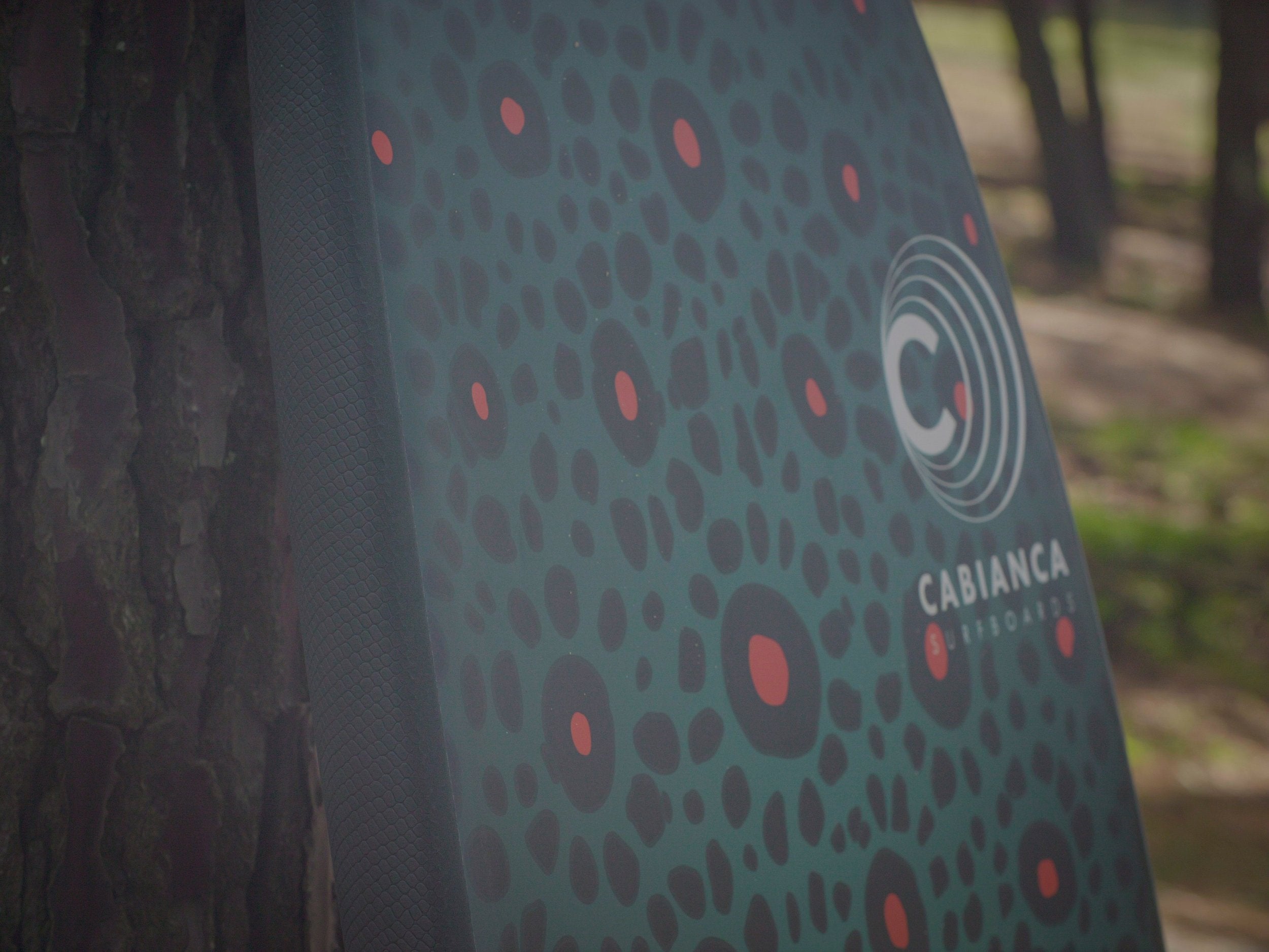 Gros plan sur une planche de surf montrant un motif léopard en vert foncé avec des points rouges. Le logo Cabianca Surfboards, composé de cercles concentriques blancs et le texte 'CABIANCA', est visible sur la surface. La texture du design est mate et professionnelle, avec un arrière-plan flou naturel