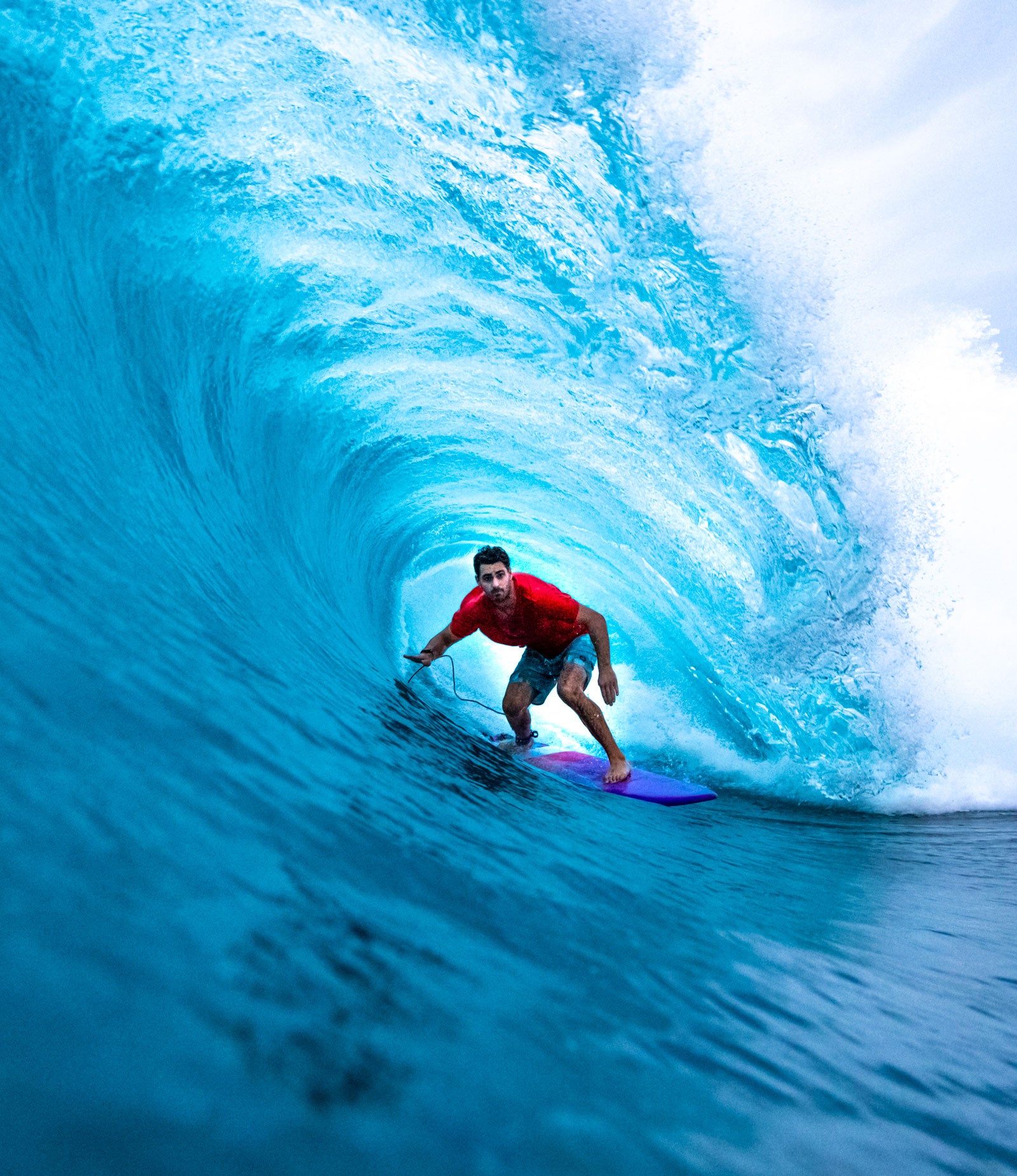Un surfeur en t-shirt rouge surfe dans un tube d'eau turquoise parfait. Il se tient en position bottom turn sur une planche bleue et violette, naviguant à l'intérieur de la vague. La photo est prise depuis l'intérieur du tube, capturant la courbe majestueuse de la vague et sa couleur bleu cristallin caractéristique des vagues tropicales. L'écume blanche couronne le haut de la vague