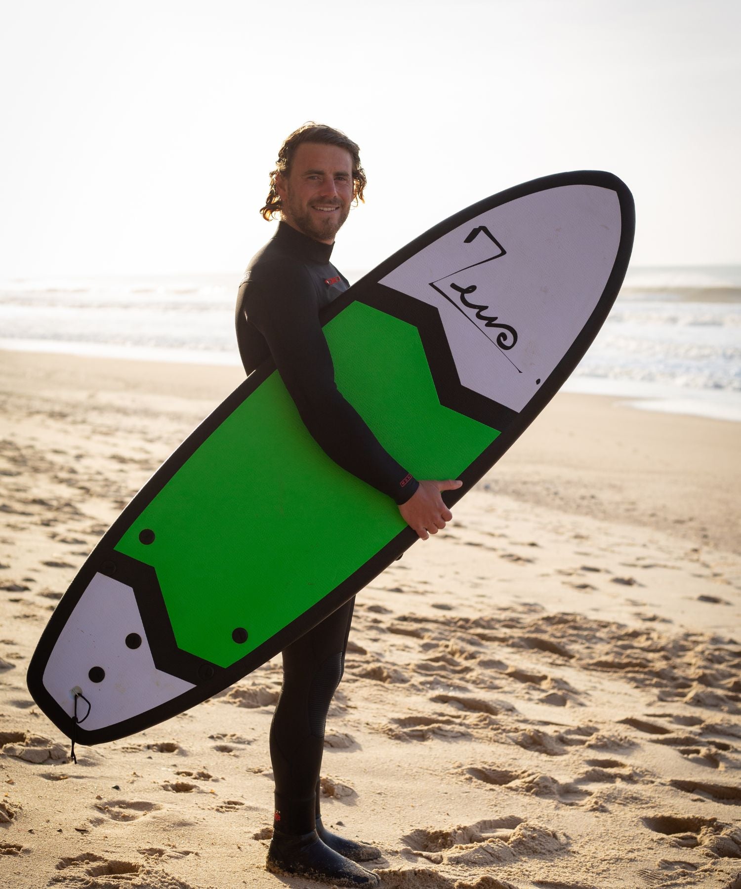 Surfeur avec une planche de surf en mousse Zeus sous le bras en train de marcher sur la plage