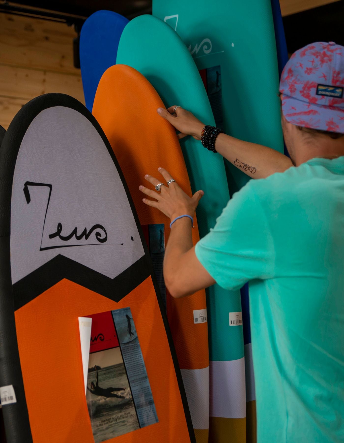 une personne en t-shirt turquoise et casquette, manipulant des planches de surf de la marque Zeus. Les planches sont rangées verticalement et présentent différentes couleurs : noir et blanc, orange, bleu et turquoise dans un surfshop