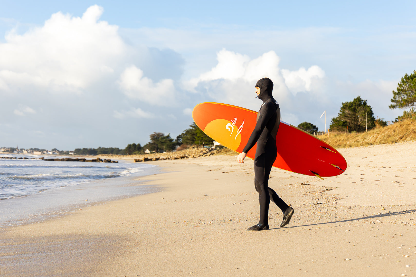 A surfer in a black wetsuit walking along a beach carrying an orange and yellow gradient surfboard. The scene shows a cloudy sky and coastal vegetation in the backgroun