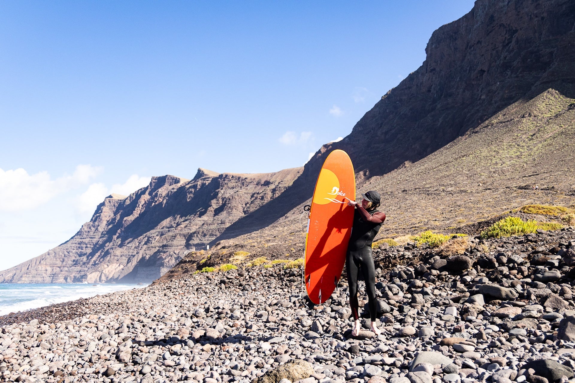 A surfer in a black wetsuit walking along a rbeach carrying an orange and yellow gradient surfboard from Zeus 