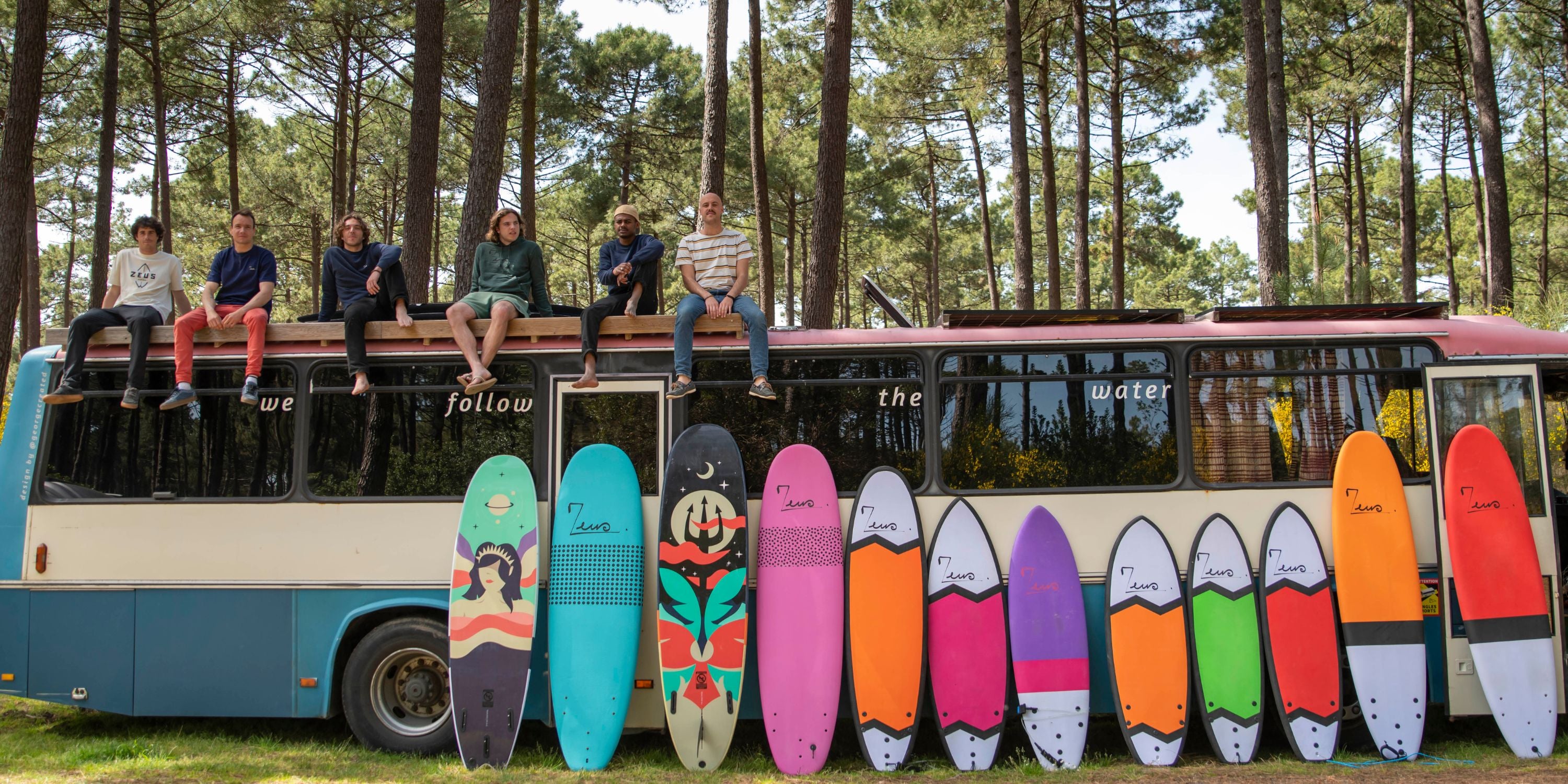  image shows a retro-style bus painted in blue and white with the text "we follow the water" written across its side. Several people are sitting casually on top of the bus's roof against a backdrop of tall pine trees. In front of the bus is a colorful display of surfboards arranged in a row - they appear to be Zeus brand surfboards in various designs and colors including mint green, turquoise, black, pink, orange, purple, and red. Each board has unique artwork and patterns