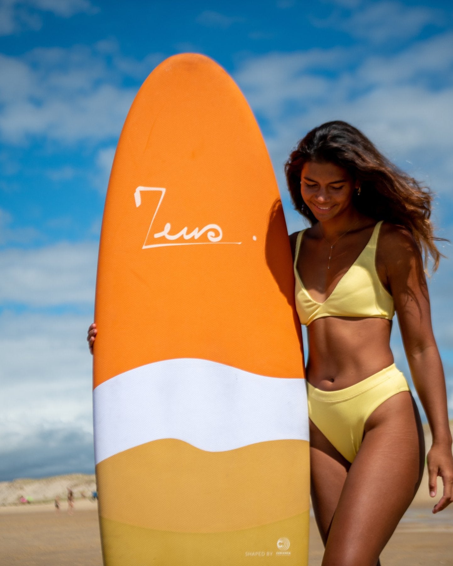 a woman in a yellow bikini holding a surfboard on the beach in France, the bord is an egg shape from Zeus 